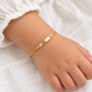 Zartes Kinderarmband aus 585 Gelbgold mit glänzender Gravurplatte, am Kinderhandgelenk zu einem weißen Oberteil getragen.