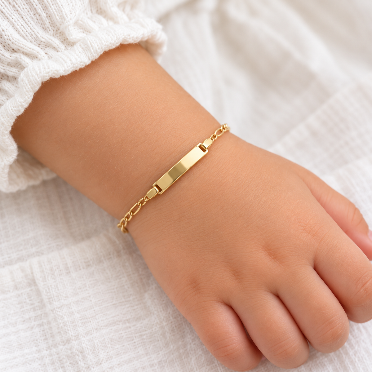 Zartes Kinderarmband aus 585 Gelbgold mit glänzender Gravurplatte, am Kinderhandgelenk zu einem weißen Oberteil getragen.